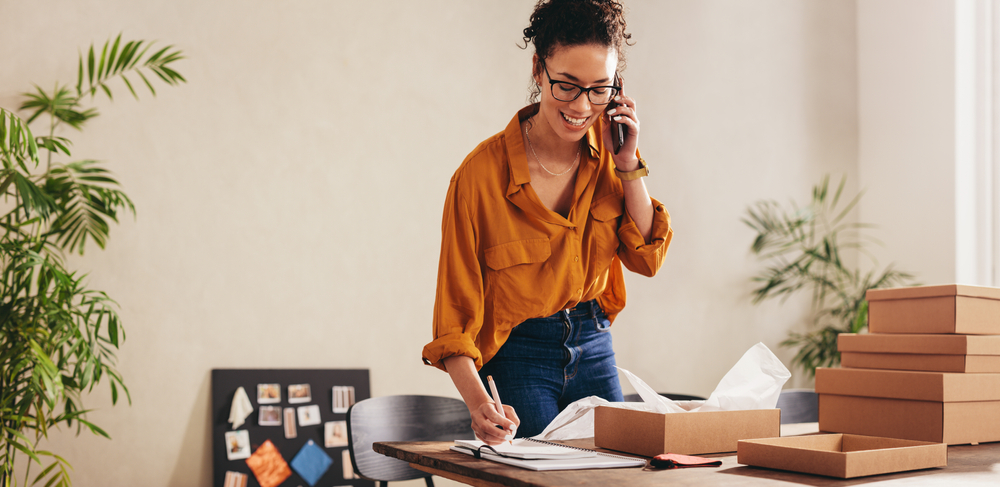 young entrepreneur woman in an orange shirt talking on the phone and taking notes on Bad Credit Merchant Account