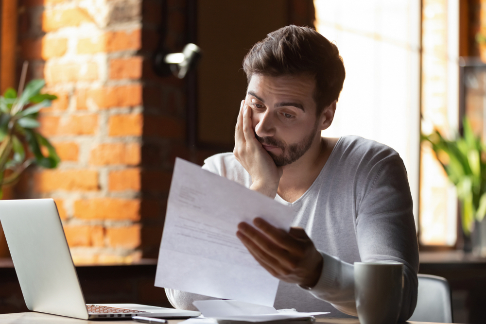 Emotional male with anxious face staring at sheet of paper sitting at a table with laptop