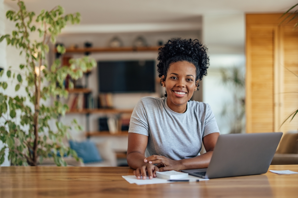 Joyful happy short haired Black business woman sitting at workplace with laptop in home office, looking at camera, smiling. Millennial worker, employee, entrepreneur head shot portrait