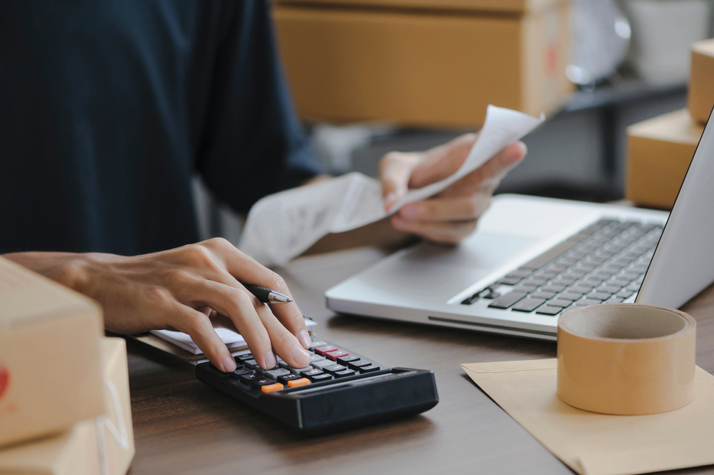 A business owner using a calculator and researching how to get a merchant account on his laptop.