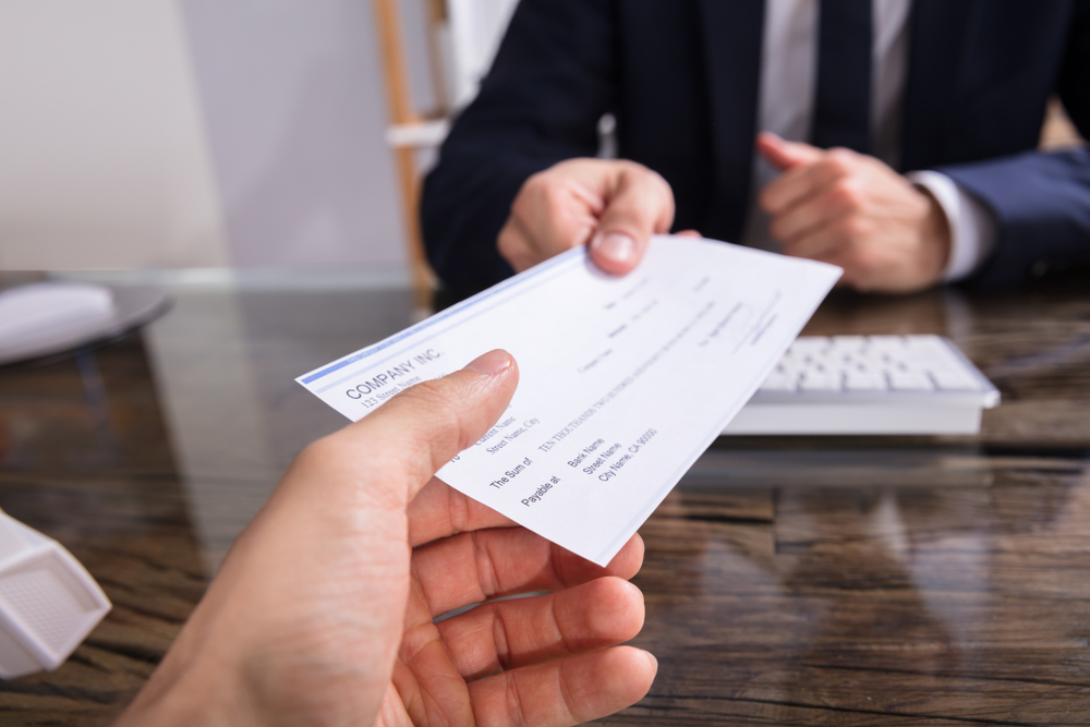 A person handing another person behind a keyboard a check that will be processed through a check processing system.
