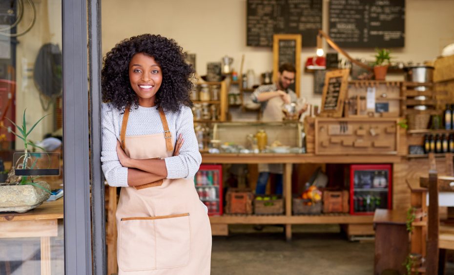 A smiling young woman wearing an apron standing by the open door of her small business.