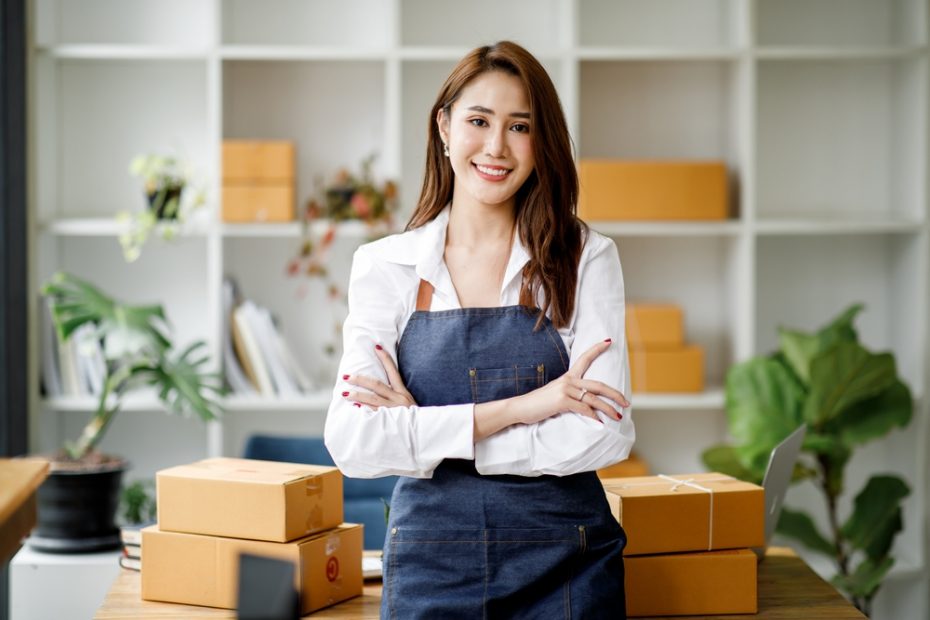 A merchant smiling and standing at her business counter surrounded by packaged orders.