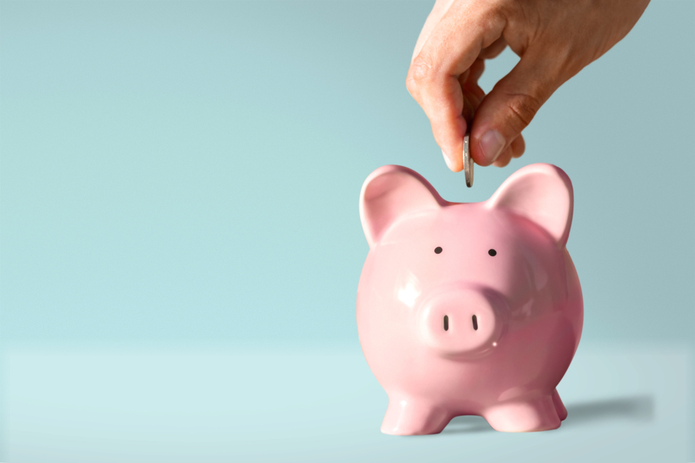 A person's hand putting coins in a piggy bank with a stack of coins on a desk, a calculator, and a pair of red-rimmed glasses.