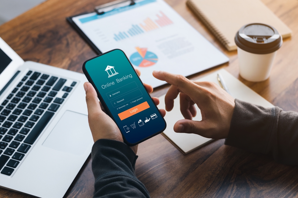 A merchant logging into their merchant account on a smartphone surrounded by a coffee, a laptop, a clipboard with financial charts, and a pen and paper.