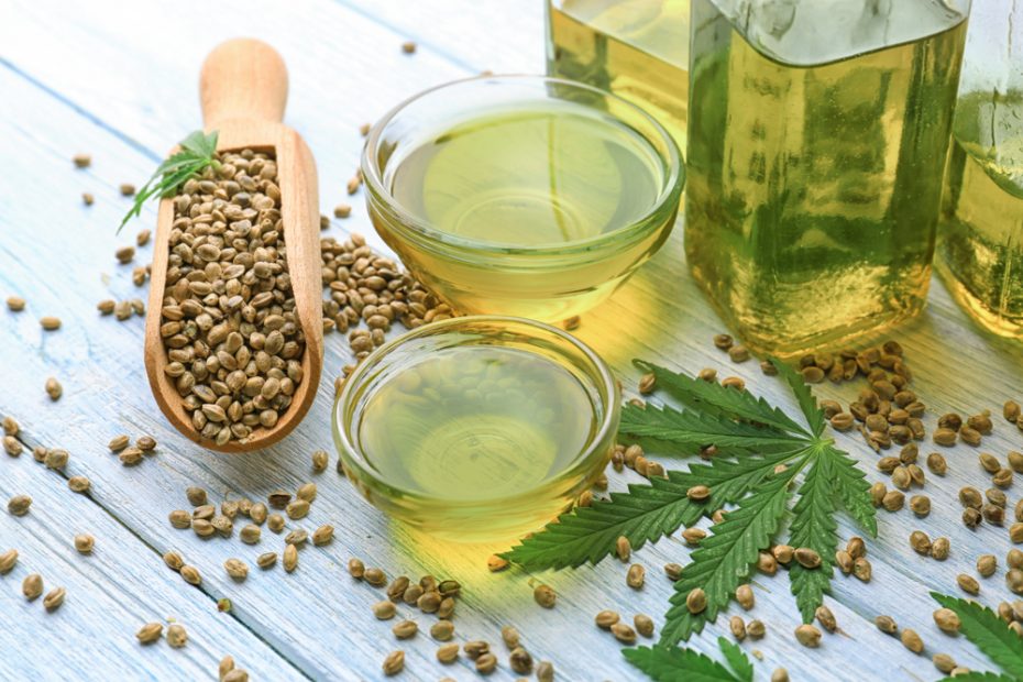 A hemp leaf surrounded by hemp seeds in a wooden scoop, two glass bowls with oil, and three glass bottles with hemp oil.