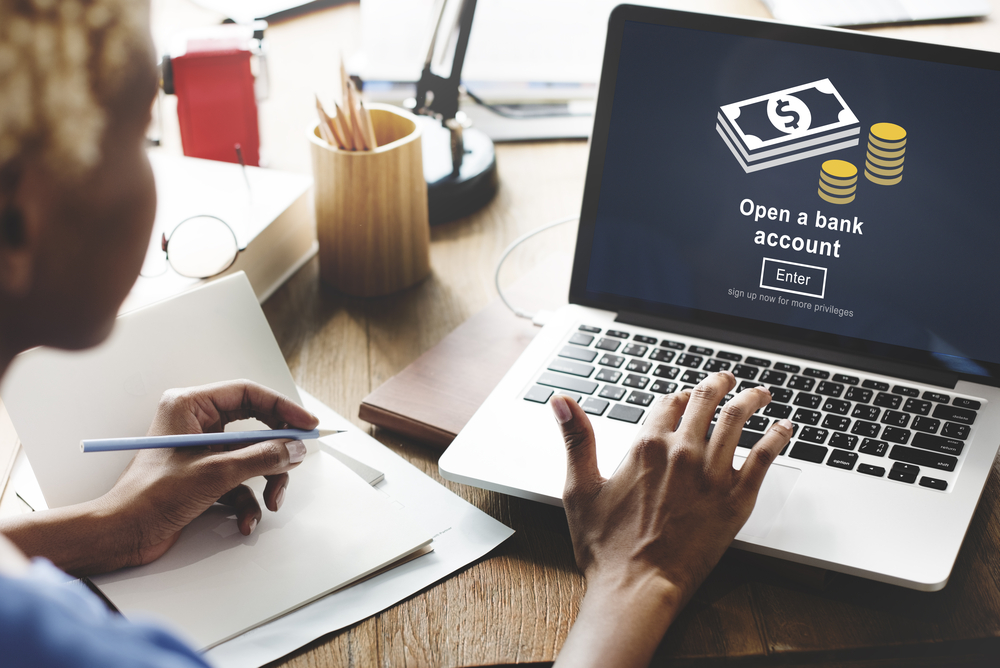 A woman on a laptop on a page to open a bank account. On the other hand, she is 
holding a pen, and a pad of paper is under her hand.