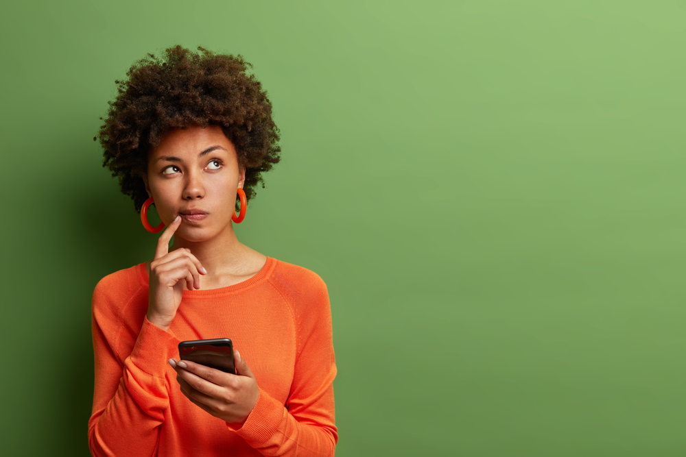 A woman in an orange shirt holding a phone and thinking about what to look for in a merchant service provider.