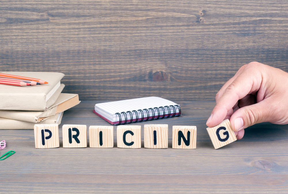 Wooden blocks spelling out the word pricing with someone placing the G block in the correct place and a notebook behind the blocks, books with colored pencils on top, and paperclips. 